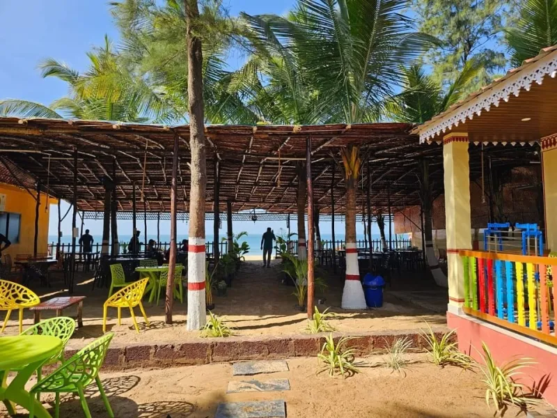 Open-air beachfront restaurant under a thatched roof, with palm trees and the sea beyond